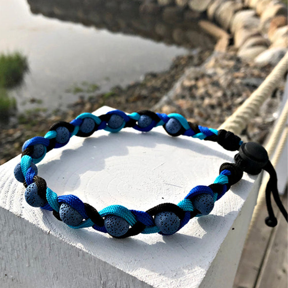 The AtlanTick Lava Collar resting on a wooden post by the water, featuring blue and black braided nylon with lava beads designed to absorb Tick Attack™ Botanical Insect Repellent. Perfect for pets and outdoor enthusiasts.