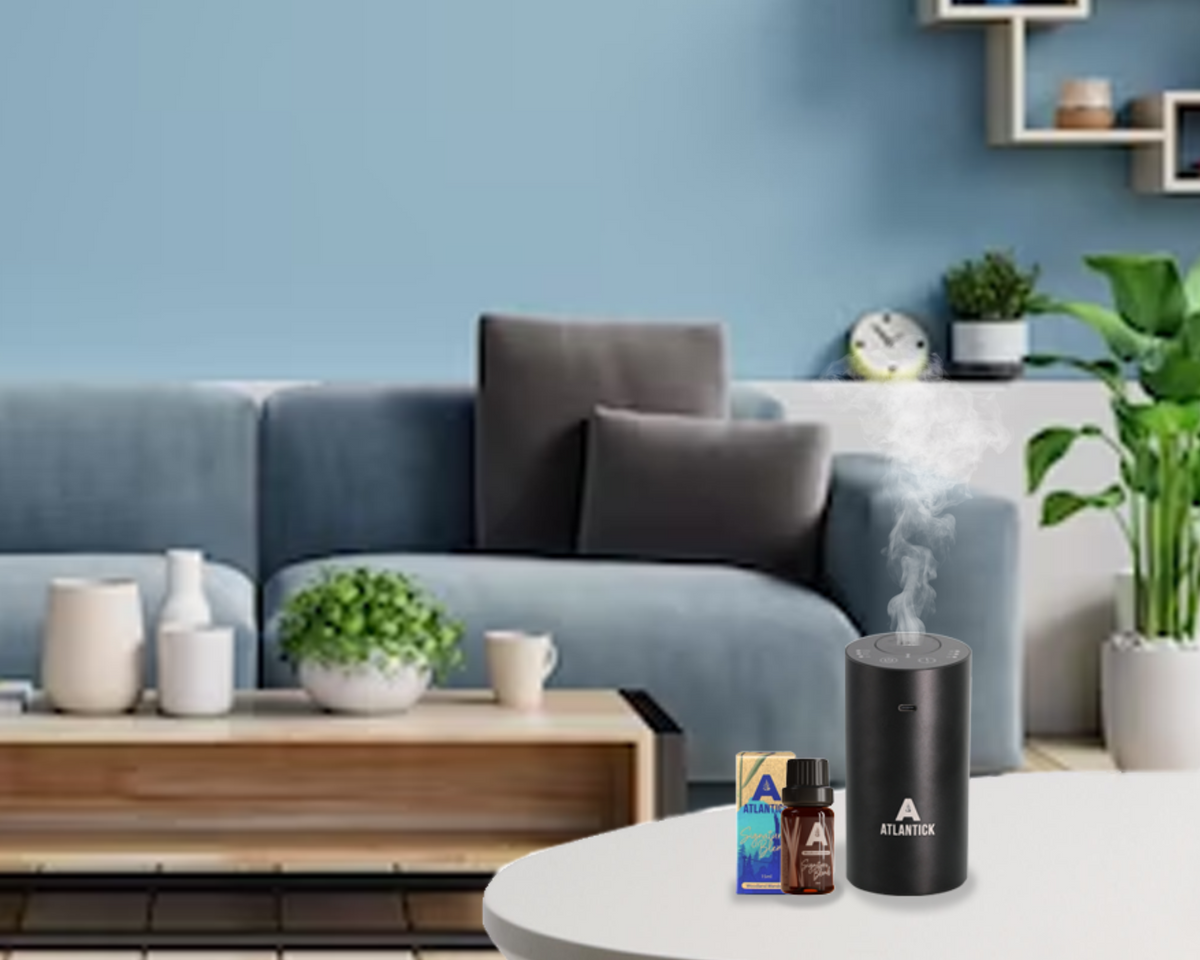 The AtlanTick Portable Air Infuser sits on a white table in a cozy, modern living room, releasing a fine mist of essential oils. Accompanied by a bottle of AtlanTick Signature Blend, the infuser blends seamlessly into the stylish space, creating a fresh, aromatic atmosphere. The neutral-toned room features a blue sofa, decorative plants, and a warm, inviting ambiance.