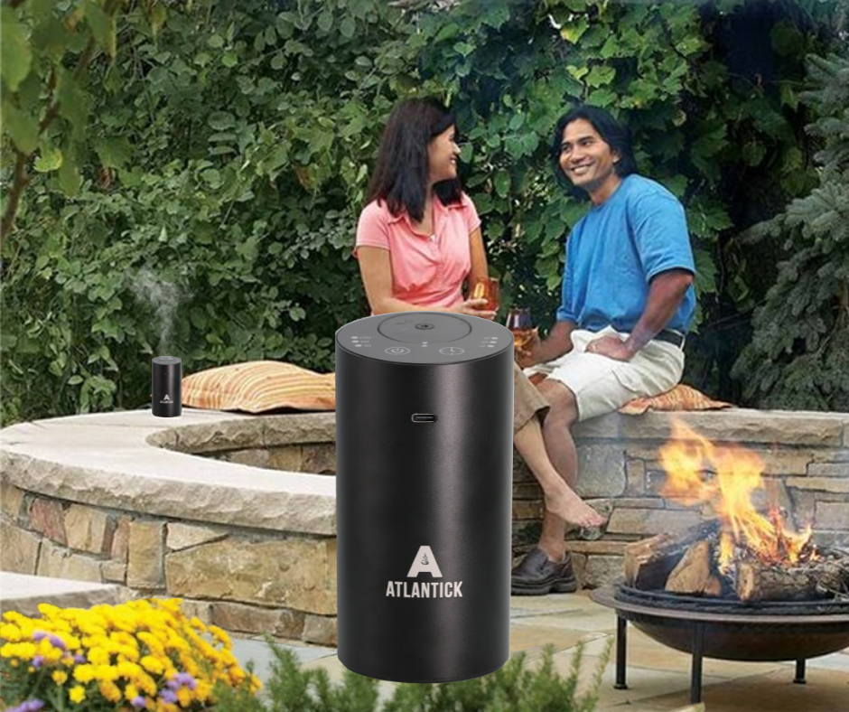 A couple enjoys a relaxing evening by an outdoor fire pit, with the AtlanTick Portable Air Infuser nearby, releasing a fine mist of essential oils. The sleek black infuser sits on a stone ledge, blending seamlessly with the natural setting. This waterless, ultrasonic diffuser enhances the outdoor atmosphere with pleasant scents while supporting a comfortable and bug-free experience.