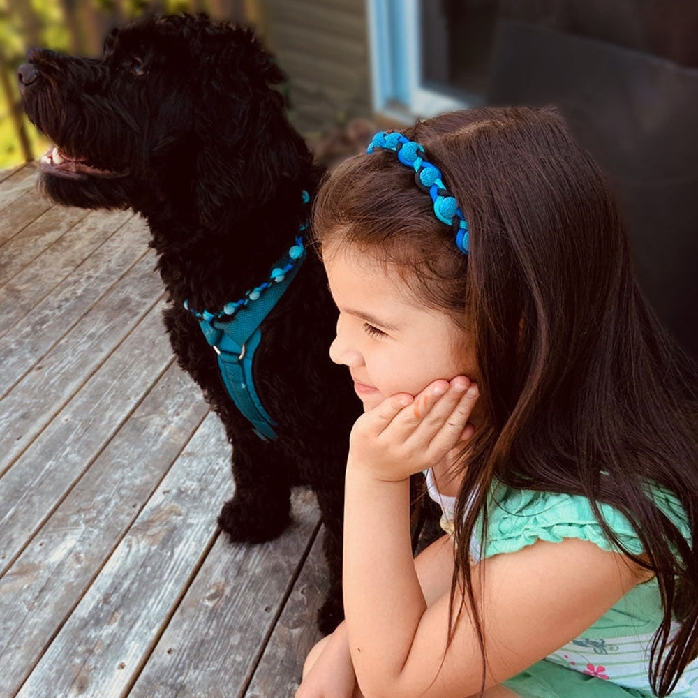 A little girl and her black dog sit together on a wooden deck, both wearing matching AtlanTick Lava Bands—one as a headband and the other as a stylish dog collar. A perfect blend of function and fashion for pets and their humans!